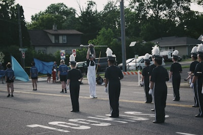 A group of marching band members stand on a street, wearing dark uniforms with white gloves and tall hats adorned with white plumes. One member in the center is dressed in white and holds up a baton. In the background, people carry large blue flags. There are some houses, parked cars, and trees along the street.