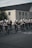 A group of marching band members is parading down a street, wearing black uniforms with white hats featuring decorative white plumes. They are playing various brass and woodwind instruments in front of a large brick building.