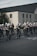 A group of marching band members is parading down a street, wearing black uniforms with white hats featuring decorative white plumes. They are playing various brass and woodwind instruments in front of a large brick building.