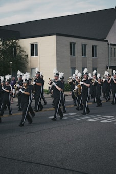 A group of marching band members is parading down a street, wearing black uniforms with white hats featuring decorative white plumes. They are playing various brass and woodwind instruments in front of a large brick building.