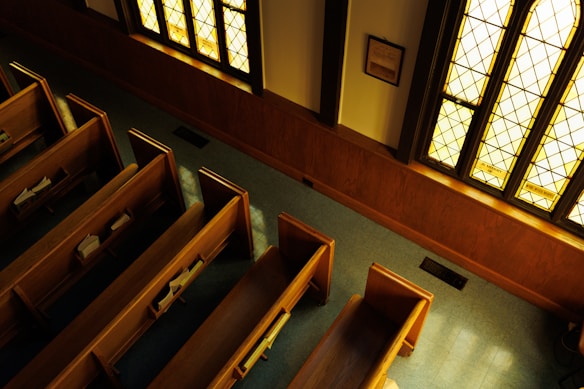 A tranquil interior view of a church, highlighting wooden pews arranged in rows. Sunlight streams through stained glass windows, casting a warm, golden glow across the scene. Hymnals are visible on some of the pews, and the room is empty, creating a serene and peaceful atmosphere.