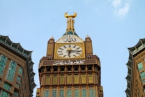 A large clock tower with a golden crescent on top, adorned with intricate designs and inscriptions in Arabic. The clock is flanked by two buildings with multiple windows, and the sky appears clear with a light blue color in the background.