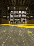 A warehouse aisle with tall metal shelves filled with stacked boxes and pallets. The perspective is low to the ground, highlighting the yellow safety lines painted on the floor. The environment seems organized and industrial, with a focus on storage and logistics.