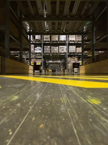A warehouse aisle with tall metal shelves filled with stacked boxes and pallets. The perspective is low to the ground, highlighting the yellow safety lines painted on the floor. The environment seems organized and industrial, with a focus on storage and logistics.