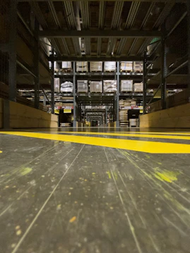 A warehouse aisle with tall metal shelves filled with stacked boxes and pallets. The perspective is low to the ground, highlighting the yellow safety lines painted on the floor. The environment seems organized and industrial, with a focus on storage and logistics.