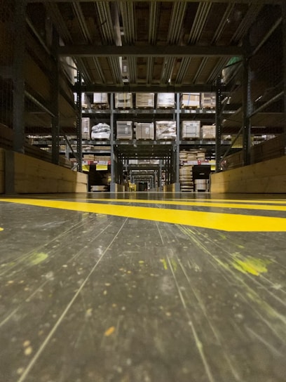 A warehouse aisle with tall metal shelves filled with stacked boxes and pallets. The perspective is low to the ground, highlighting the yellow safety lines painted on the floor. The environment seems organized and industrial, with a focus on storage and logistics.