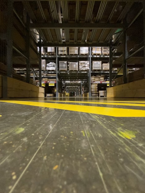 A warehouse aisle with tall metal shelves filled with stacked boxes and pallets. The perspective is low to the ground, highlighting the yellow safety lines painted on the floor. The environment seems organized and industrial, with a focus on storage and logistics.