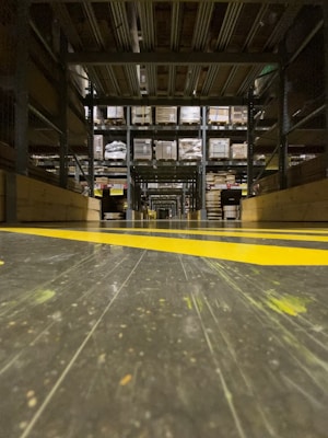 A warehouse aisle with tall metal shelves filled with stacked boxes and pallets. The perspective is low to the ground, highlighting the yellow safety lines painted on the floor. The environment seems organized and industrial, with a focus on storage and logistics.