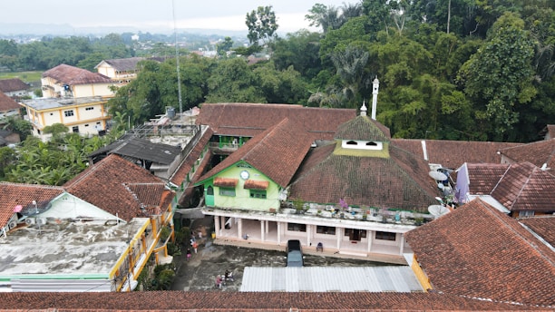 Aerial view of a village with buildings featuring red-tiled roofs, surrounded by dense green trees and vegetation. In the center, a building with a distinct green facade and a small dome stands out. People and a vehicle are visible in the courtyard area. Various structures and rooftops are seen in the vicinity, indicating a residential neighborhood.