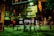 An urban street scene at night featuring a vintage-style hotel building with illuminated windows and a prominent sign. Pedestrians are seen walking past, their motion creating a blurred effect. The street is dimly lit with a combination of streetlights and neon signage, providing a moody, cinematic atmosphere.