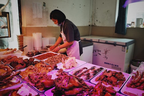 A woman wearing a dark shirt and apron is working in what appears to be a food market or a kitchen. She is handling food items placed on trays in front of her, which include various kinds of meat and sausages. The room has a freezer in the background and stacks of disposable containers to the left.