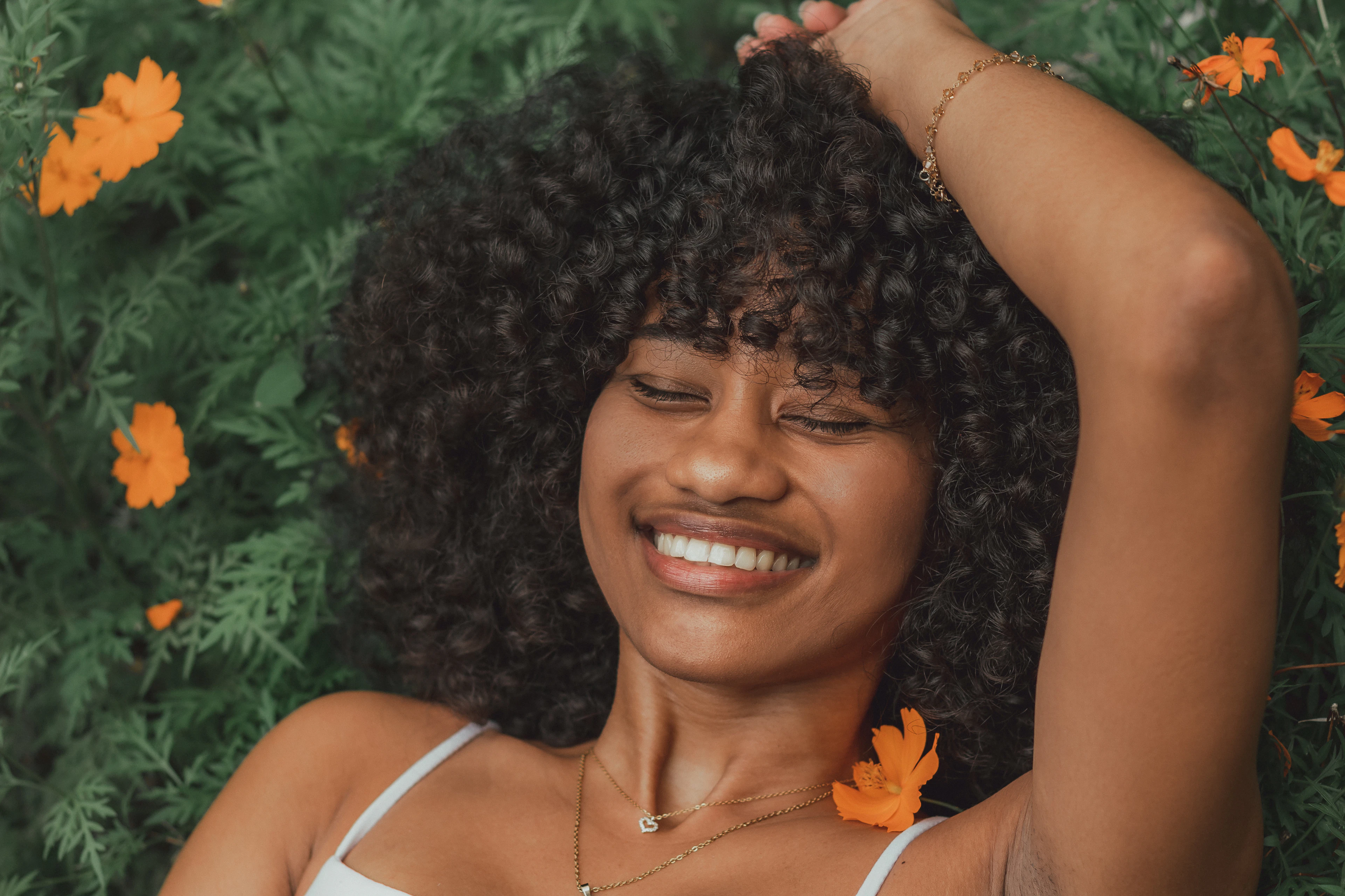 Woman with full, defined curls smiling with her eyes closed, flowers behind her
