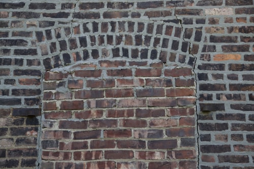 A section of a brick wall with an arch shape that has been filled in with mismatched bricks. The colors vary from dark gray to reddish-brown, and there are visible cracks in the structure.