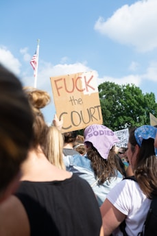 A group of people is gathered outdoors, holding signs with various messages. Prominent among the signs is one that reads 'FUCK the COURT' in bold letters. The group appears to be participating in a protest or demonstration.