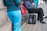 A person stands against a wall wearing blue athletic leggings with a geometric pattern. They are carrying a red bag over their shoulder. In the background, a seated person in a dark jacket and black pants is visible on the sidewalk, holding a shopping bag.