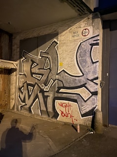 Graffiti art with bold, angular designs is painted on a wall which has a slightly worn appearance. A shadow of a person holding a spray can is visible on the ground, indicating recent graffiti activity. There is a height restriction sign indicating 'Max Høyde 3.25m'. The phrase 'Night Paint' is graffitied in red near the bottom. A can is resting on the ground near the wall.