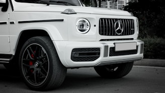 A close-up view of the front side of a luxury white SUV with a prominent circular headlight and the brand's logo in the center of the grille. The vehicle features black alloy wheels with red brake calipers, and it is parked on a smooth surface.