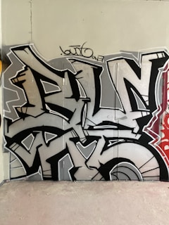 A wall is covered with expressive graffiti composed of bold, angular designs in black, white, and some red tones. The artwork features intricate lines and letters with a dynamic and urban style, typical of street art.