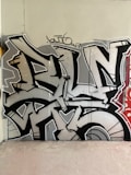 A wall is covered with expressive graffiti composed of bold, angular designs in black, white, and some red tones. The artwork features intricate lines and letters with a dynamic and urban style, typical of street art.