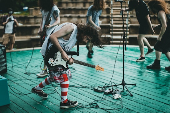 A group of musicians perform energetically on a stage with wooden planks. The central figure is playing an electric guitar while bending forward with dynamic movement. Other band members are in the background, adding to the lively scene. The venue seems to be an outdoor amphitheater with tiered seating.