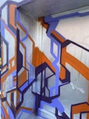 Geometric graffiti artwork featuring interconnected angular shapes in vibrant colors on a textured surface. The design cleverly uses layers and shadows to create a 3D effect, set against a grey metallic background.