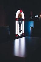 A vintage jukebox illuminated against a dark background, glowing with white and red lights. The setting appears to be dimly lit, adding a nostalgic and retro vibe.