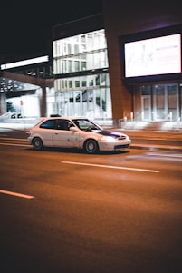 A white car is driving at night on a multi-lane road in an urban area, with modern buildings illuminated in the background. The road is empty except for the car, and the streetlights cast a warm glow on the asphalt. A large digital billboard is visible on one of the buildings.