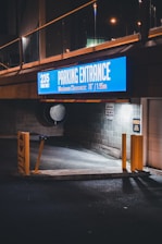 An illuminated sign displaying 'Parking Entrance 235, Maximum Clearance: 78" / 1.95m' hangs above a dimly lit parking entrance. Yellow barriers and stop signs are positioned in front of the entrance. The scene is captured at night, casting shadows and reflections on the pavement.