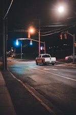 A police vehicle is seen parked at an intersection at night under the glow of streetlights. The street is mostly empty with a few other cars observed in the background. The traffic lights are illuminated, emphasizing red and green signals. The surroundings appear quiet and calm.