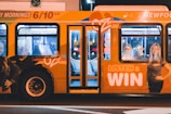 A brightly colored bus with promotional graphics parked at night under artificial lighting. The bus features large images of smiling people and text promoting a radio station with the slogan 'LISTEN & WIN'. The interior is partially visible through the windows.