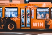 A brightly colored bus with promotional graphics parked at night under artificial lighting. The bus features large images of smiling people and text promoting a radio station with the slogan 'LISTEN & WIN'. The interior is partially visible through the windows.