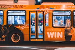 A brightly colored bus with promotional graphics parked at night under artificial lighting. The bus features large images of smiling people and text promoting a radio station with the slogan 'LISTEN & WIN'. The interior is partially visible through the windows.
