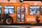 A brightly colored bus with promotional graphics parked at night under artificial lighting. The bus features large images of smiling people and text promoting a radio station with the slogan 'LISTEN & WIN'. The interior is partially visible through the windows.
