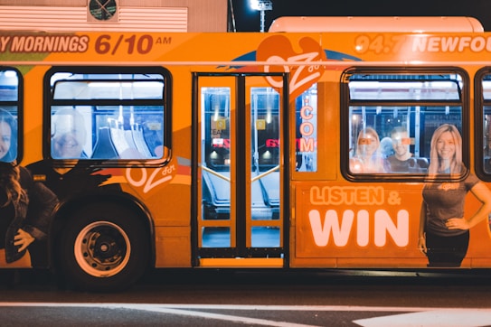 A brightly colored bus with promotional graphics parked at night under artificial lighting. The bus features large images of smiling people and text promoting a radio station with the slogan 'LISTEN & WIN'. The interior is partially visible through the windows.