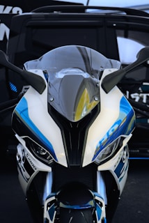 A close-up view of the front of a sports motorcycle, featuring a sleek design with aerodynamic lines and a prominent windshield. The motorcycle is predominantly white with blue and black accents, including branding and decals. The background shows part of a car or van, which is slightly out of focus.