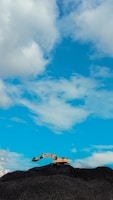 A construction excavator is positioned at the peak of a large mound of dark gravel or earth, set against a bright blue sky with scattered white clouds.