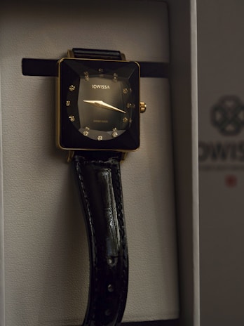 A luxurious wristwatch with a square gold-toned case, adorned with sparkling diamond-like hour markers. The watch face displays minimalistic hands and the brand name Jowissa. A glossy black leather strap complements the elegant design, housed in a sophisticated presentation box.