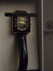 A luxurious wristwatch with a square gold-toned case, adorned with sparkling diamond-like hour markers. The watch face displays minimalistic hands and the brand name Jowissa. A glossy black leather strap complements the elegant design, housed in a sophisticated presentation box.