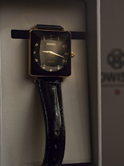 A luxurious wristwatch with a square gold-toned case, adorned with sparkling diamond-like hour markers. The watch face displays minimalistic hands and the brand name Jowissa. A glossy black leather strap complements the elegant design, housed in a sophisticated presentation box.