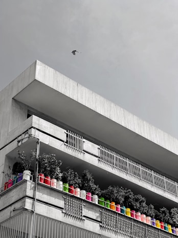 A modern concrete building with two levels of balconies, each lined with vibrant, multicolored potted plants. A single bird is flying in the sky above the building.