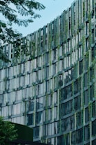 A modern building fa&ccedil;ade featuring a grid-like pattern with vertical and horizontal beams. The structure incorporates greenery, with plants interspersed between the window panels, creating an eco-friendly design. The glass windows reflect light and sky, while trees frame the image, emphasizing a blend of nature and architecture.