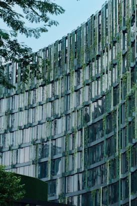 A modern building façade featuring a grid-like pattern with vertical and horizontal beams. The structure incorporates greenery, with plants interspersed between the window panels, creating an eco-friendly design. The glass windows reflect light and sky, while trees frame the image, emphasizing a blend of nature and architecture.