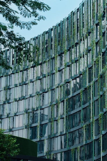 A modern building fa&ccedil;ade featuring a grid-like pattern with vertical and horizontal beams. The structure incorporates greenery, with plants interspersed between the window panels, creating an eco-friendly design. The glass windows reflect light and sky, while trees frame the image, emphasizing a blend of nature and architecture.