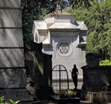 A stone mausoleum with intricate architectural details, including a prominent window featuring a six-pointed star design. The structure is surrounded by lush green foliage and several gravestones. A silhouette of a statue or figure stands near the entryway.