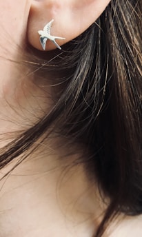 A close-up of an ear with a silver bird-shaped earring. Strands of brown hair are visible around the ear.
