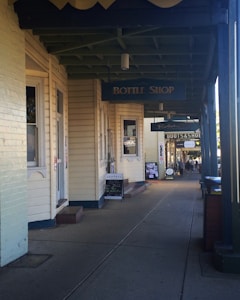 A street view of a series of shops with vintage architecture, featuring wooden siding and overhanging signs. There are signs for a bottle shop, a cafe, and a boutique. The pathway is lined with pillars and has a relaxed, small-town feel.