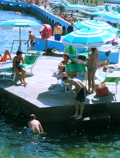 A lively scene at a beach resort with people relaxing and sunbathing under green and white umbrellas. Some individuals are lounging on green chairs while others are standing or swimming near the shore. The setting exudes a summery atmosphere with clear blue water and a sunny sky. A red lifebuoy is mounted on the blue wall nearby.