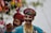Two individuals dressed in vibrant traditional clothing with intricate beadwork and embroidery, adorned with colorful turbans and face paint. Both appear to be engaged in a cultural or festive activity, as one holds a decorated stick with tassels. The background is blurred with hints of greenery.