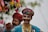 Two individuals dressed in vibrant traditional clothing with intricate beadwork and embroidery, adorned with colorful turbans and face paint. Both appear to be engaged in a cultural or festive activity, as one holds a decorated stick with tassels. The background is blurred with hints of greenery.