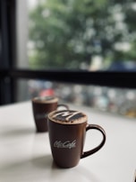 Two brown coffee mugs with a logo sit on a white surface. The mugs are topped with a frothy layer, likely featuring cocoa powder art. In the background, there is a blurred view of green foliage and a window, suggesting an indoor cafe setting.